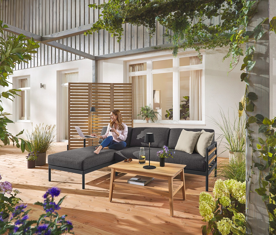 Žena sedí na prémiové zahradní rohové sedací soupravě »Elin« s flexibilním paravánem v pozadí. Vidět je také černá venkovní dobíjecí stojací a stolní lampa LED a zahradní konferenční stolek.