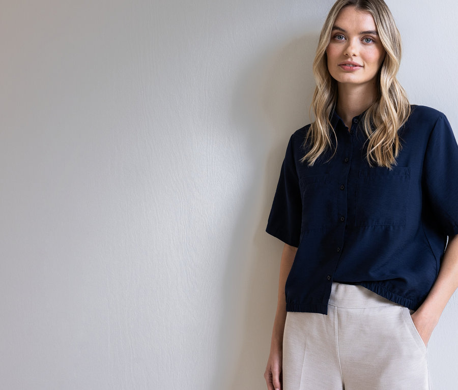 Žena s blond vlasy v tmavě modré košili a béžových kalhotách pózuje před světle šedou stěnou.