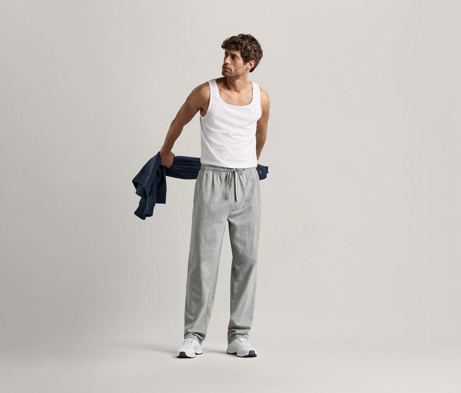 Muž v nátělníku a šedých pohodlných kalhotách drží na rameni modrou košili.