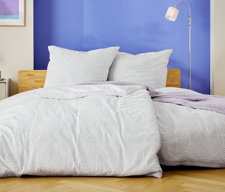 Postel s bílým povlečením s geometrickým vzorem a dvěma polštáři stojí před modrou zdí.