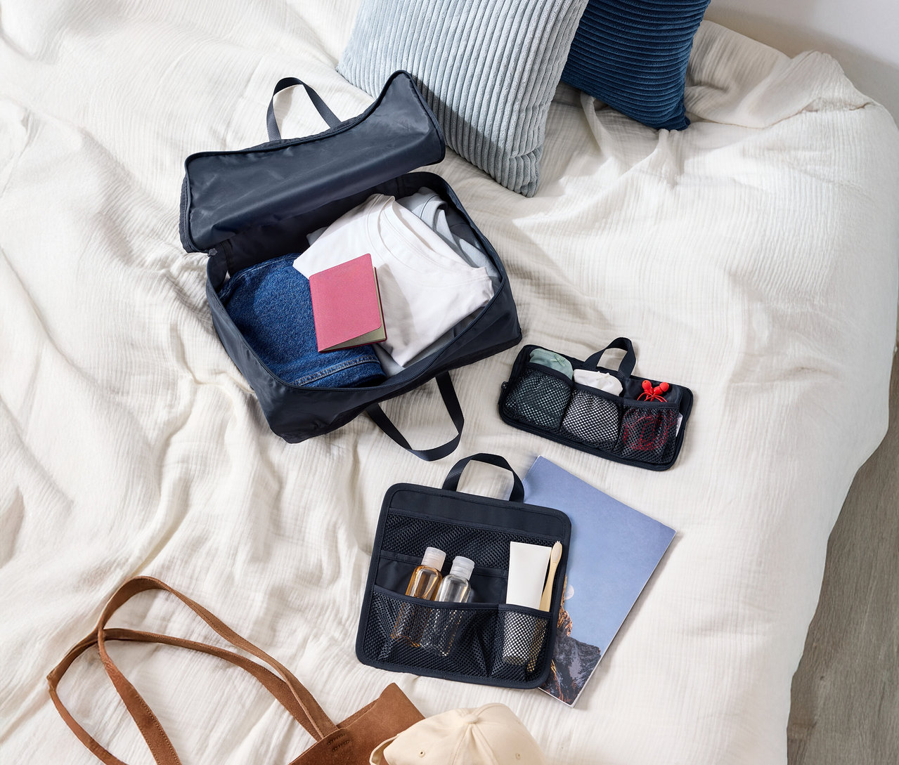 Pohled shora na kufr s oblečením, dva polštáře, kosmetickou taštičku, kabelku a čepici na posteli.