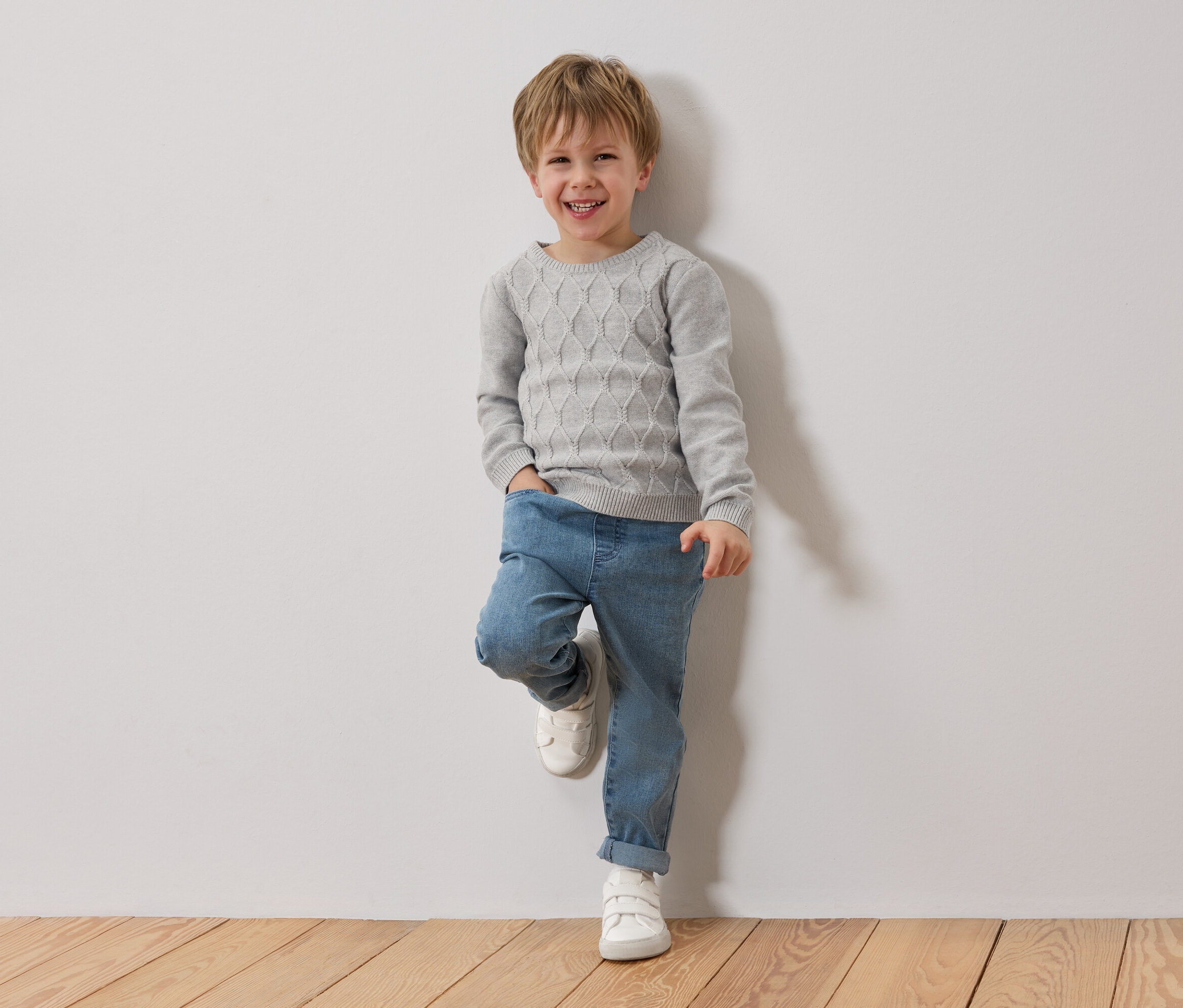 Usmívající se blonďatý chlapec se opírá o bílou zeď. Má na sobě šedý svetr, modré džíny a bílé boty.