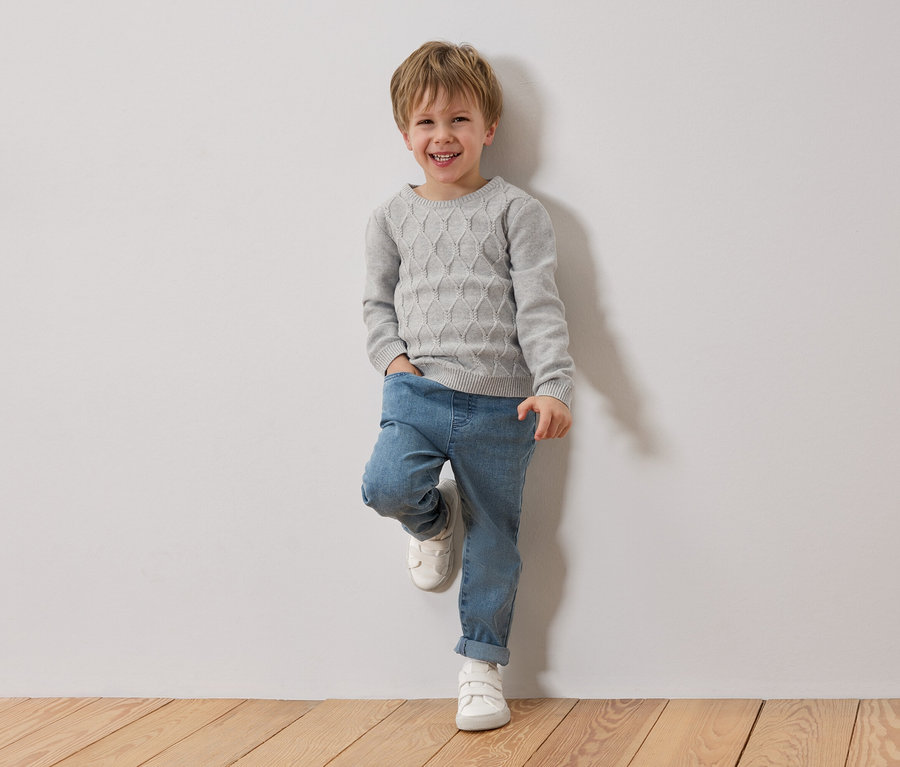 Usmívající se blonďatý chlapec se opírá o bílou zeď. Má na sobě šedý svetr, modré džíny a bílé boty.