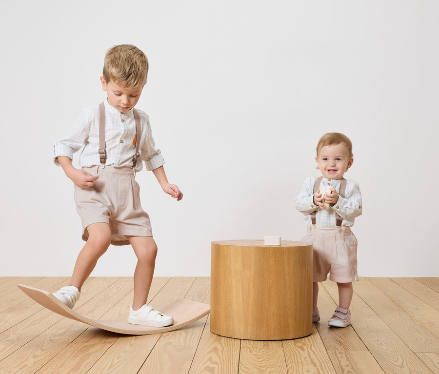 Dvě děti, chlapec balancuje na dřevěné desce, kojenec stojí vedle dřevěného stolku a drží hračku.