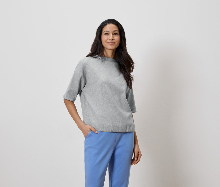 Žena s tmavými vlasy má na sobě šedý top a modré kalhoty, ruka v kapse.