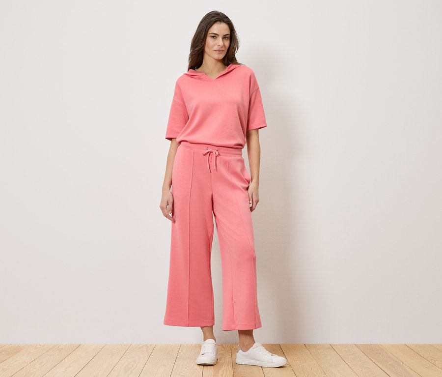 Žena stojí před zdí a má na sobě korálový dvoudílný outfit skládající se z mikiny s krátkým rukávem a culottes kalhot spolu s bílými teniskami.