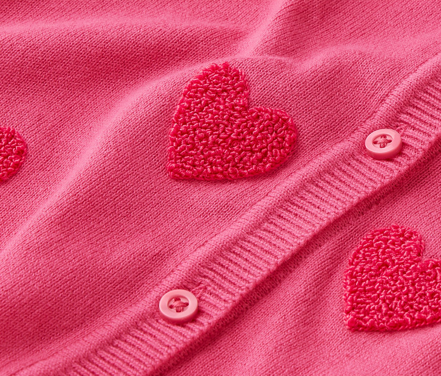 Detailní záběr na růžový kardigan s aplikacemi ve tvaru srdce a knoflíky.