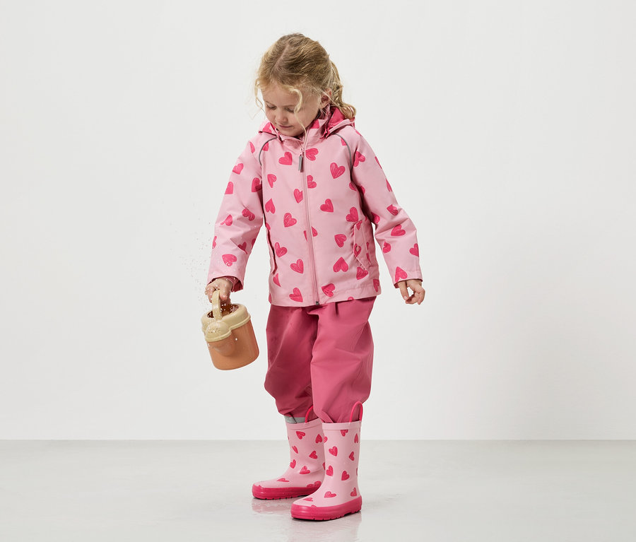 Holčička sype písek z hračkového kbelíku, oblečená do růžové bundy se vzorem srdíček, sladěných kalhot a holínek.