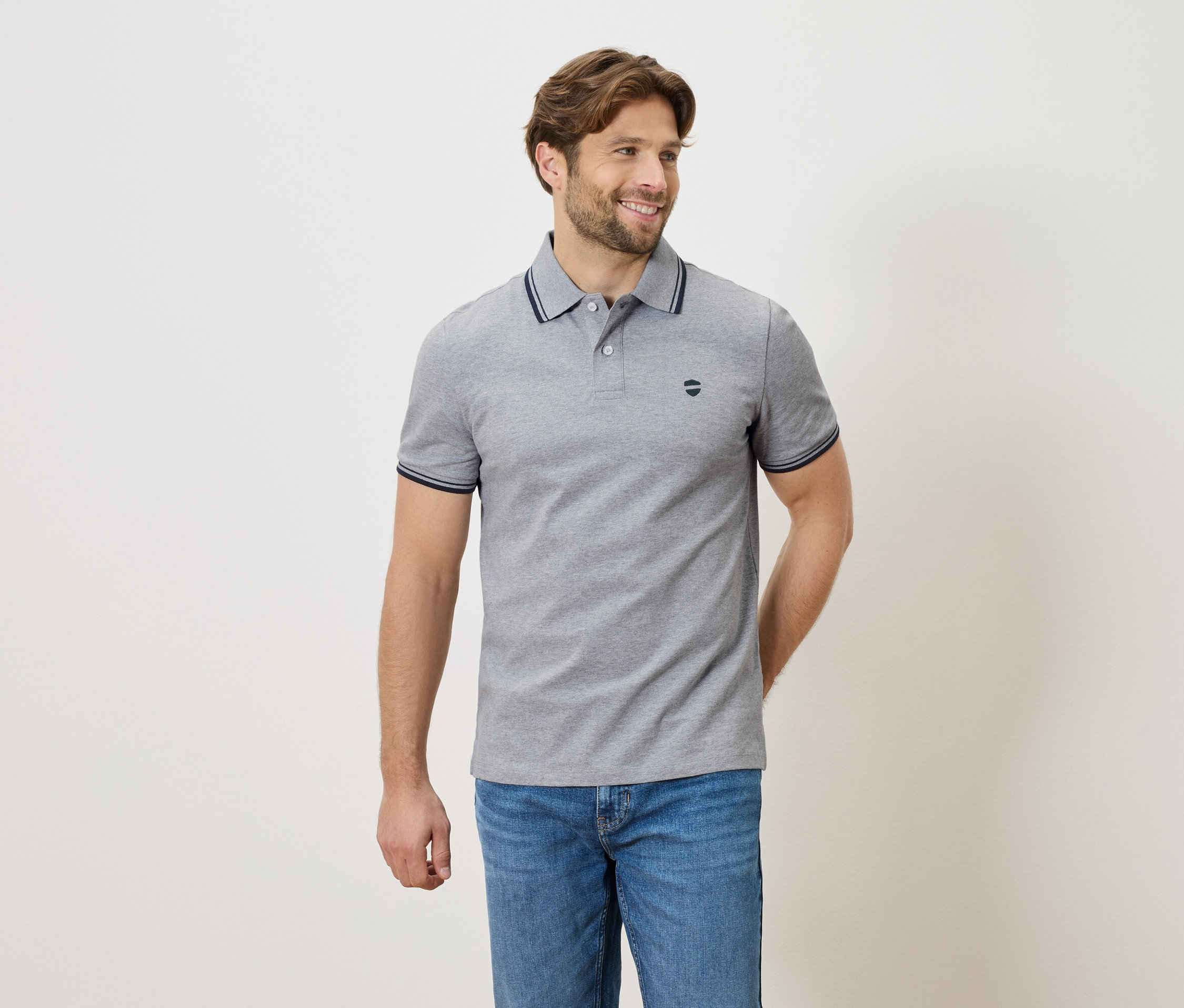 Muž v šedém polo tričku a džínách stojí na neutrálním pozadí.