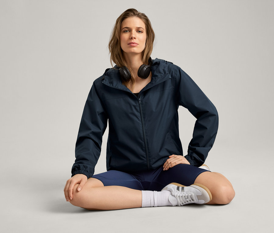 Žena sedí v tureckém sedu a má na sobě bundu do deště, sluchátka, šortky a tenisky.