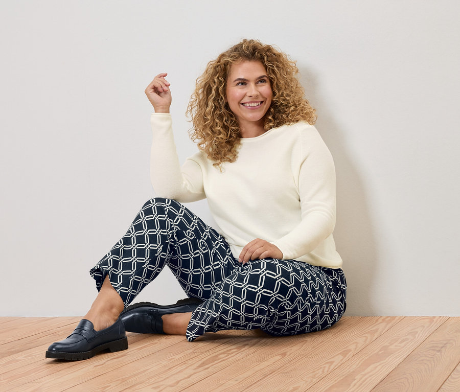 Žena s kudrnatými vlasy sedí na dřevěné podlaze, má na sobě krémový svetr a modré kalhoty s geometrickými vzory.