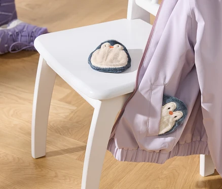 Světle fialová bunda visí na bílé židli, s plyšovou hračkou tučňáka na sedadle a další v kapse bundy. Dvoje fialové dětské boty jsou na dřevěné podlaze.