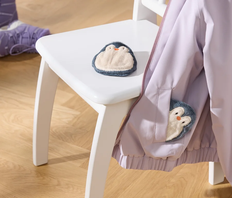 Světle fialová bunda visí na bílé židli, s plyšovou hračkou tučňáka na sedadle a další v kapse bundy. Dvoje fialové dětské boty jsou na dřevěné podlaze.