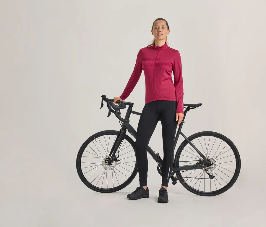 Žena stojí vedle černého silničního kola, má na sobě fuchsiový cyklistický dres s dlouhým rukávem, černé cyklistické kalhoty a černé boty.