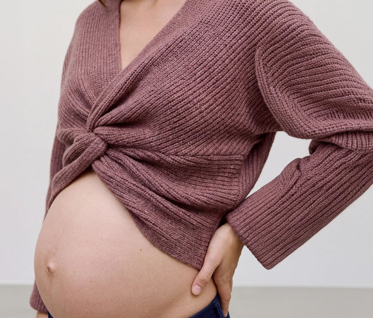 Těhotná žena má na sobě růžový zkrácený pletený top MAMALICIOUS »SVALA« s překříženou přední částí, který odhaluje její břicho. Její levá ruka spočívá na boku.