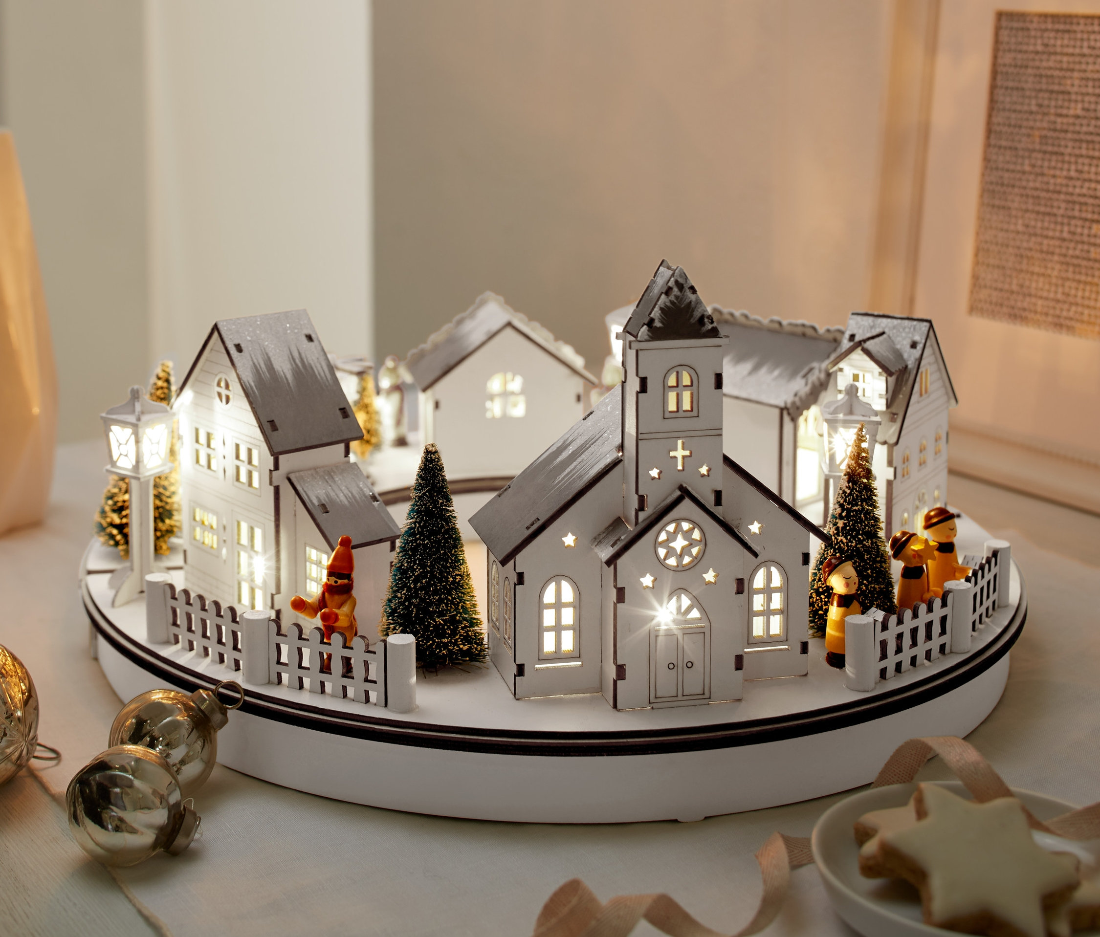 Osvětlená dřevěná vánoční vesnička s LED světly, vedle vázy, ozdoby a talíře sušenek.