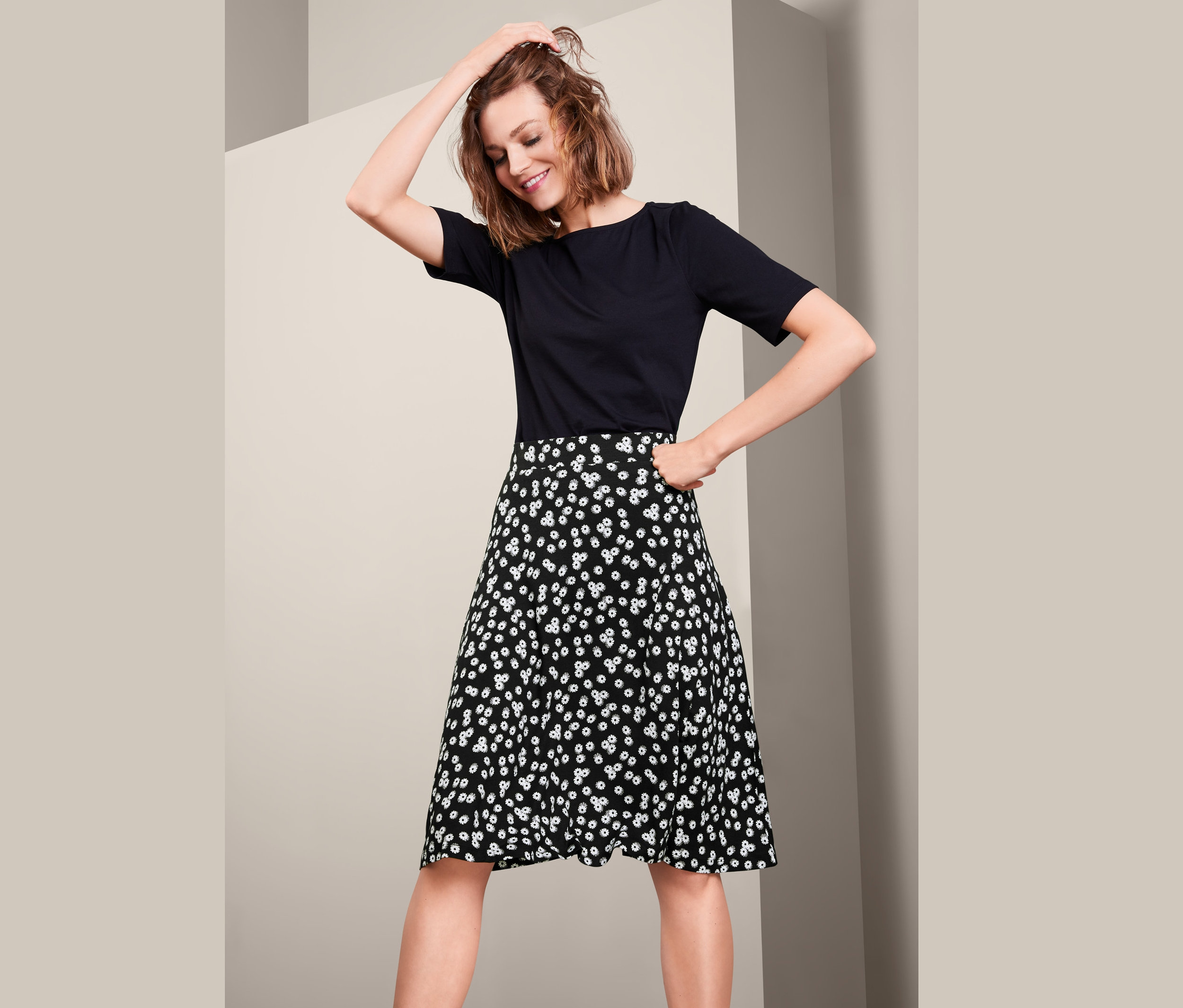 Žena pózuje v černém tričku a žerzejové sukni s kopretinovým vzorem.