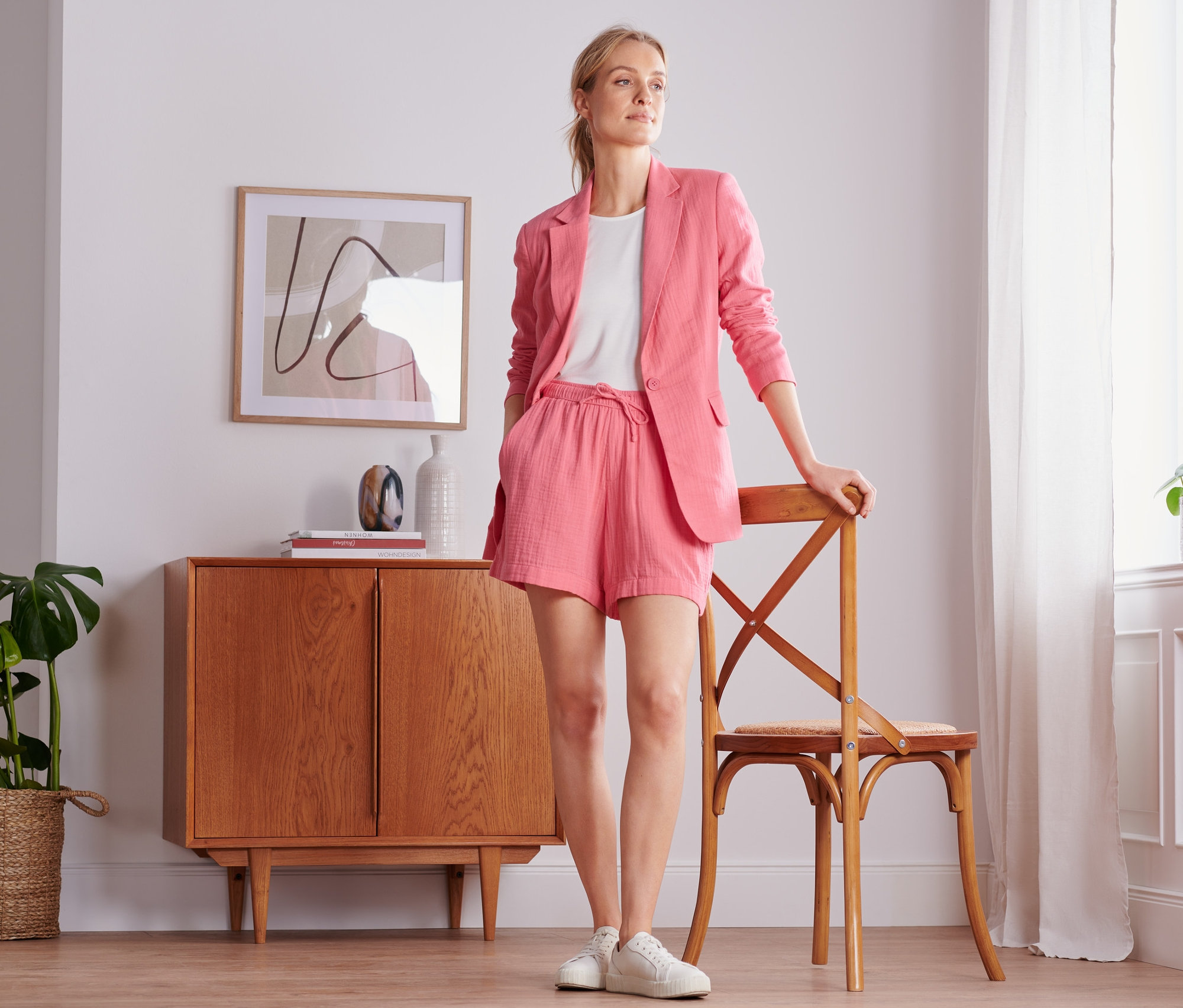 Žena pózuje v korálovém mušelínovém saku a šortkách. Opírá se o židli vedle okna.