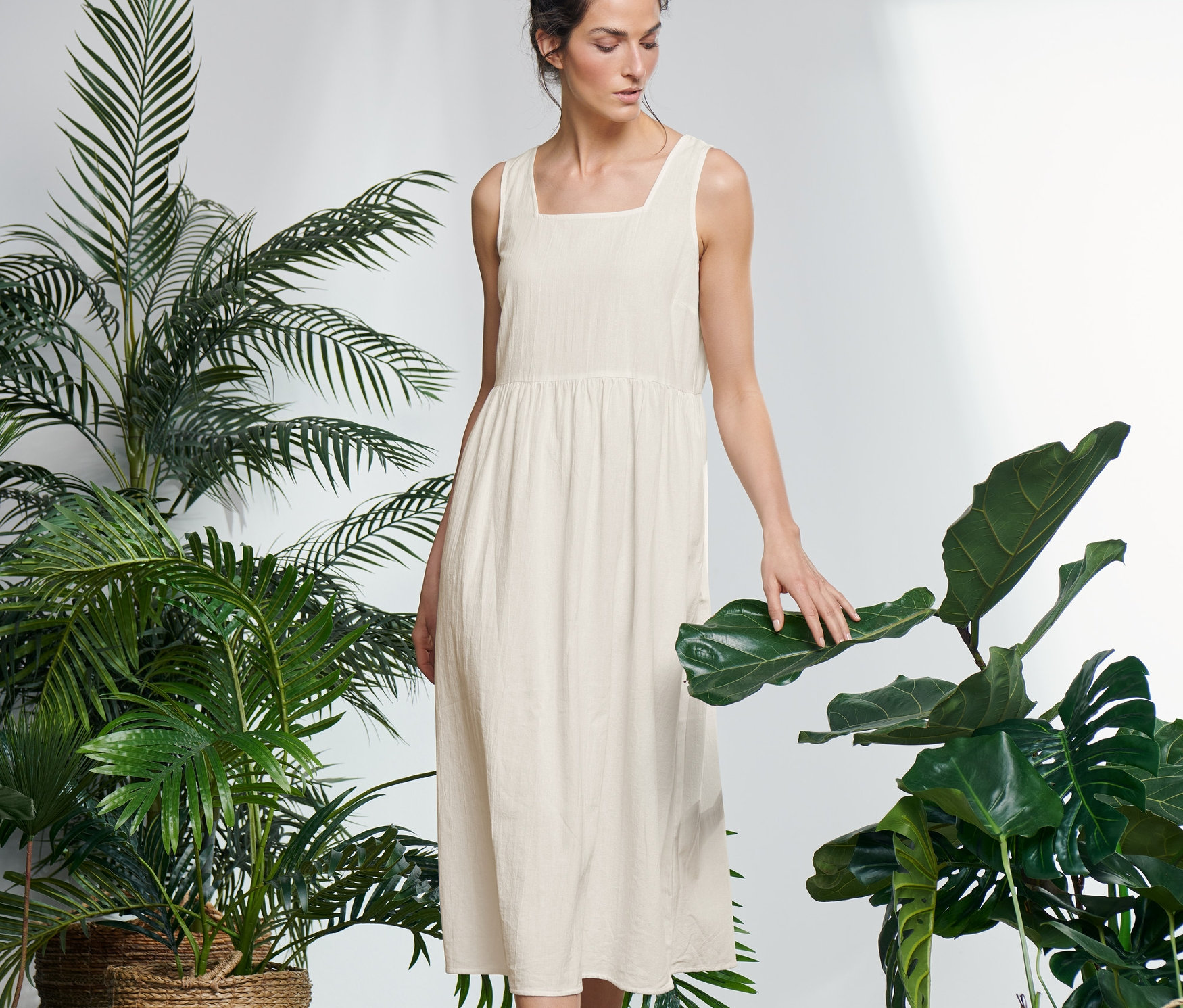Žena stojí v místnosti s rostlinami a dotýká se listu. Má na sobě midi šaty se širokými ramínky a tenisky.
