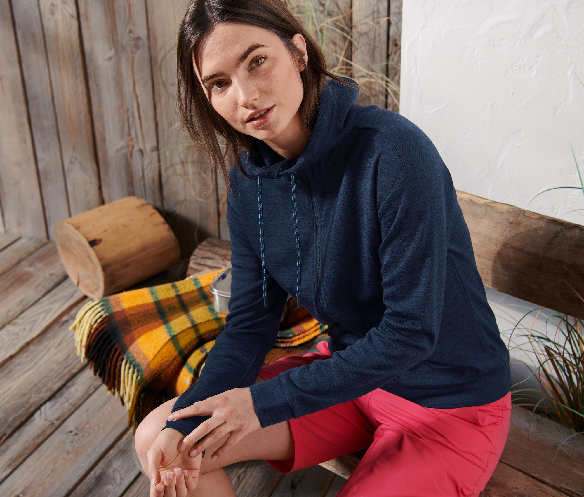 Žena sedí na dřevěné lavičce a má na sobě tmavě modrou mikinu s kapucí z merino vlny a růžové šortky.
