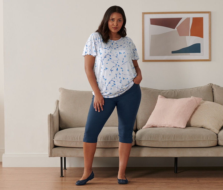 Žena stojí v místnosti a má na sobě bílé halenkové triko s netopýřími rukávy, tmavě modré tříčtvrteční kalhoty z bengalínu a kožené baleríny.