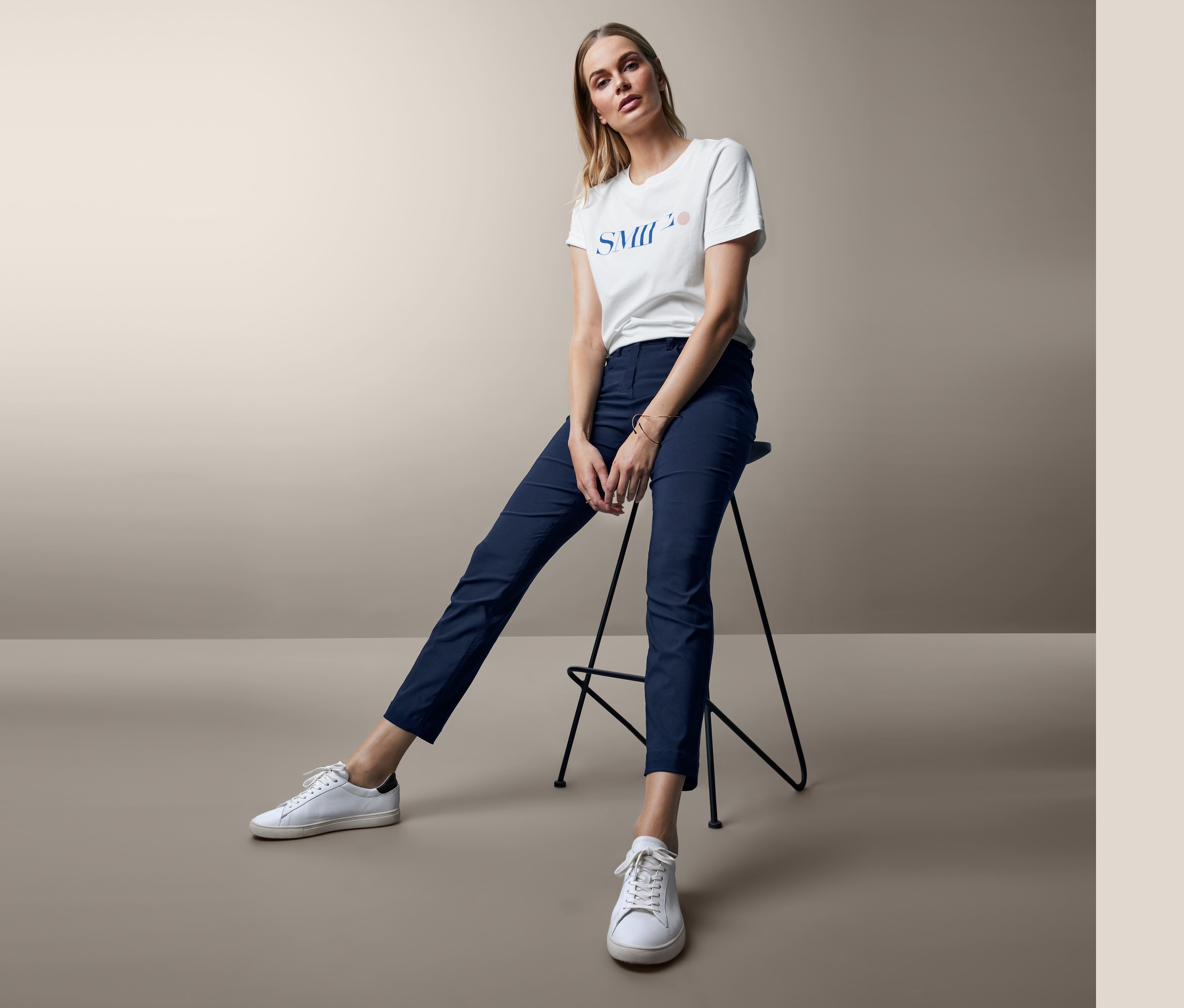 Žena sedí na stoličce a má na sobě bílé tričko a tmavě modré bengalínové kalhoty.