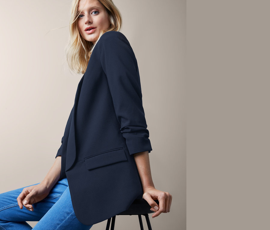 Blonďatá žena sedí a má na sobě blejzr s nařasenými rukávy a džíny.