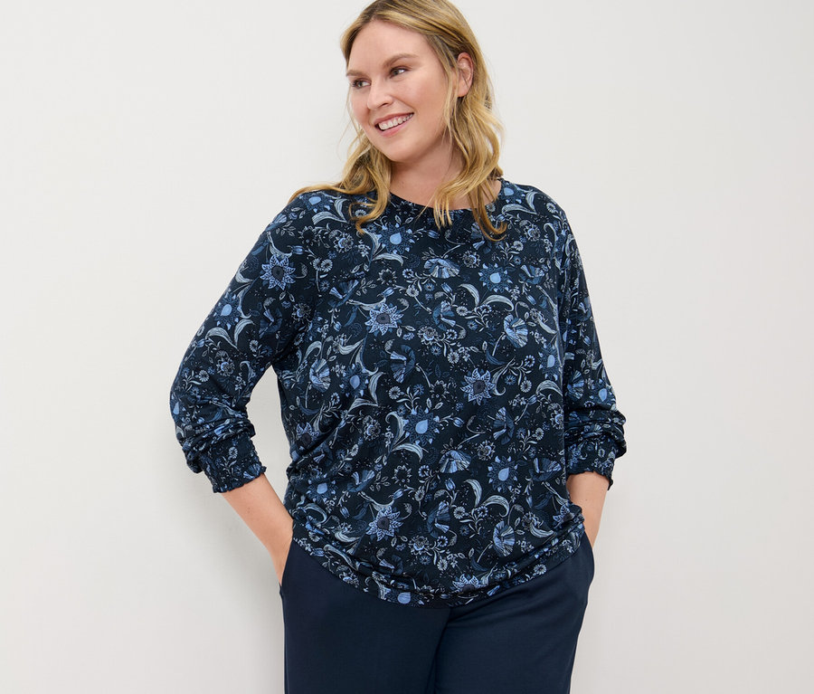 Žena pózuje v halenkovém triku s potiskem a pohodlných žerzejových kalhotách.