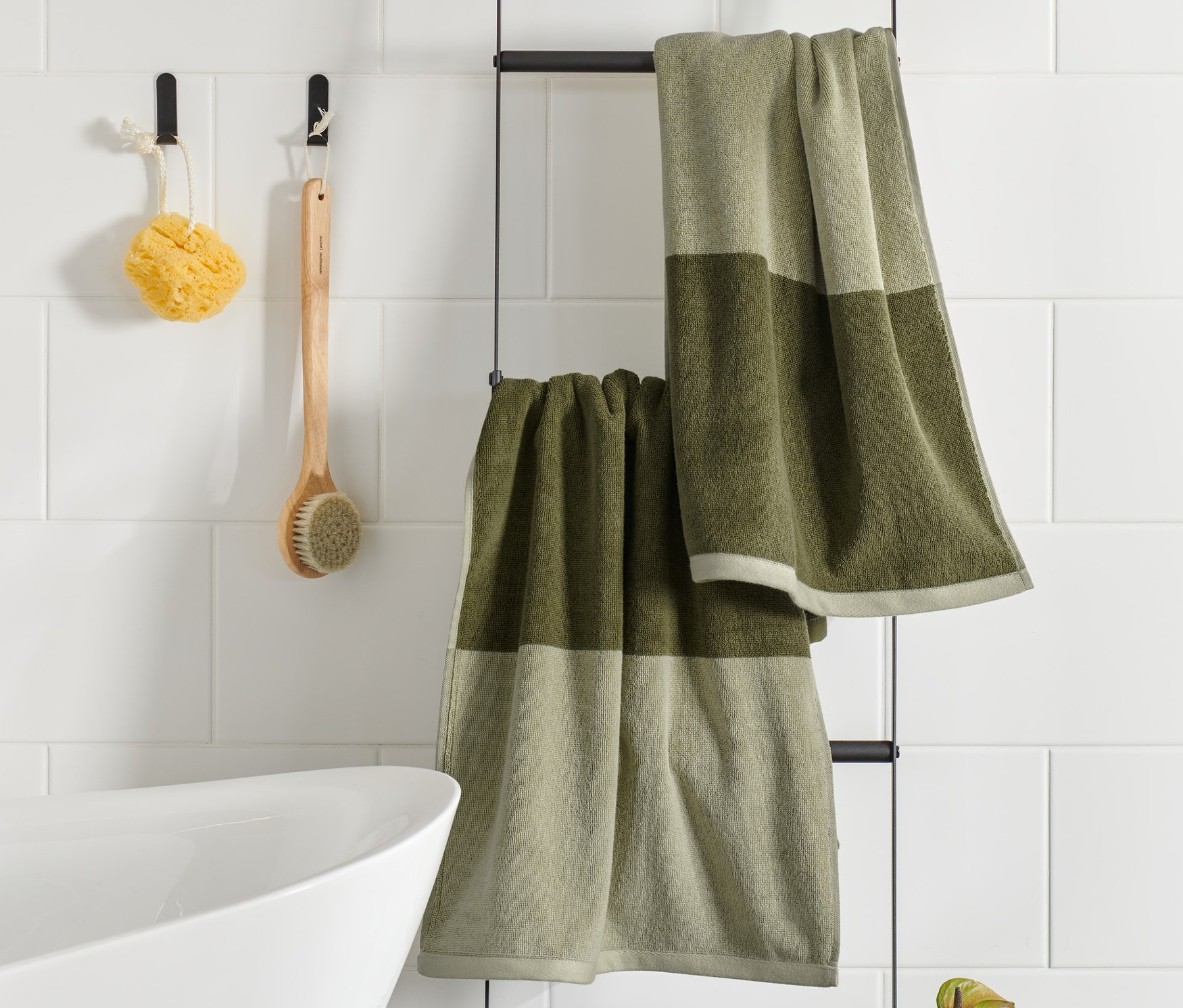 Dva vysoce kvalitní žakárové ručníky v pískově a mechově zelené barvě visí na černém rámu v koupelně.