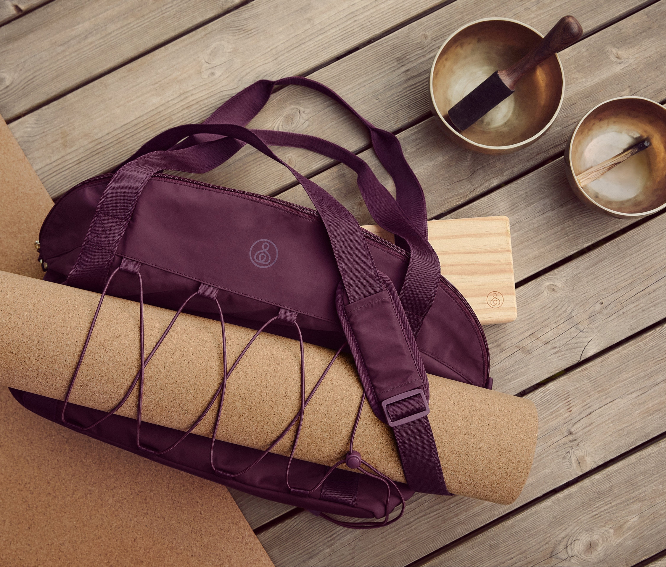 Na dřevěné podlaze leží korková podložka na jógu, sportovní taška, blok na jógu a dvě tibetské mísy.