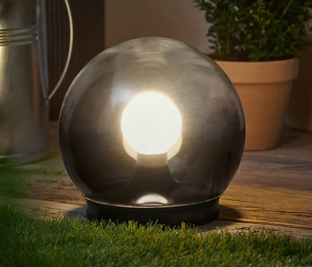 Kulovitá lampa z kouřového skla stojí na dřevěném povrchu vedle rostlin.