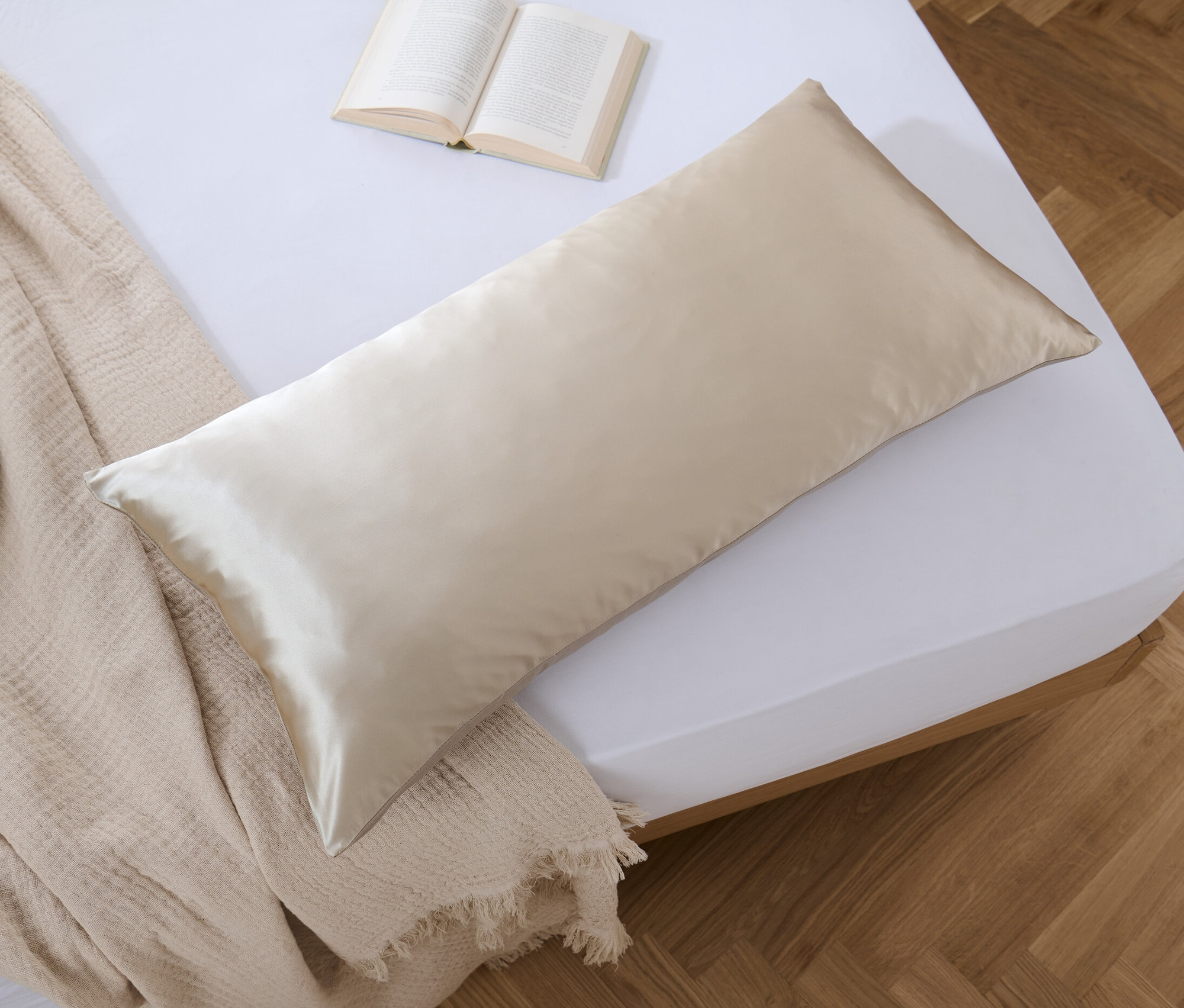 Béžový hedvábný povlak na polštář o rozměrech 100 x 65 cm leží na posteli vedle otevřené knihy a přehozu.