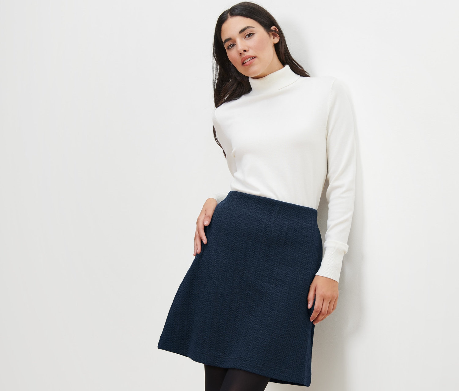 Žena pózuje v teplákové sukni s copánkovým vzorem a bílém roláku.