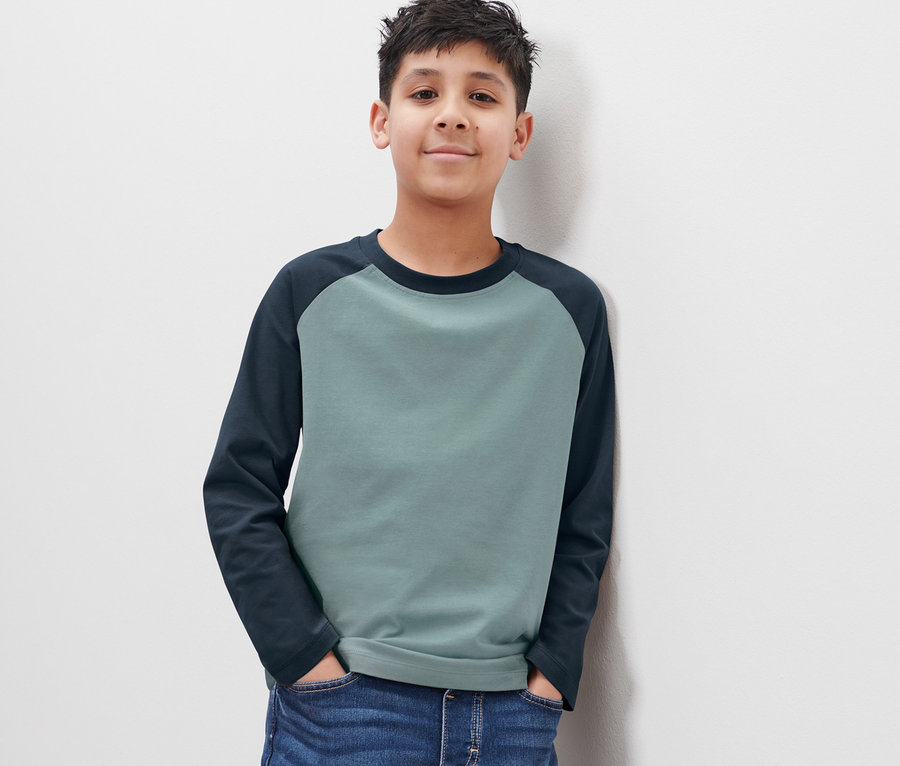 Chlapec v modrých džínách a dvoutónovém tričku s dlouhým rukávem stojí zády k bílé zdi.