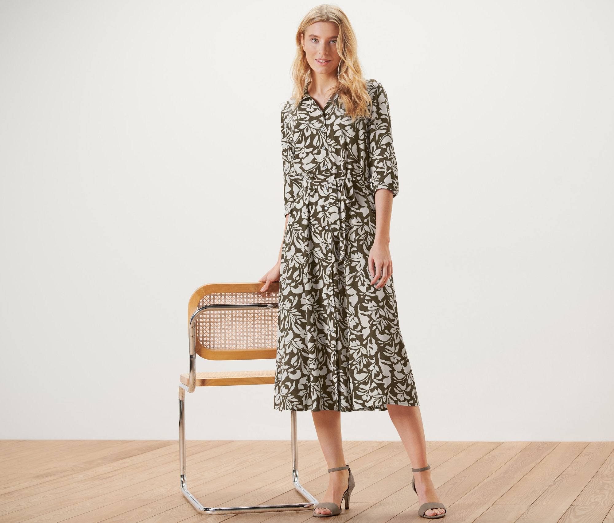 Blonďatá žena pózuje vedle židle a má na sobě halenkové šaty s potiskem.