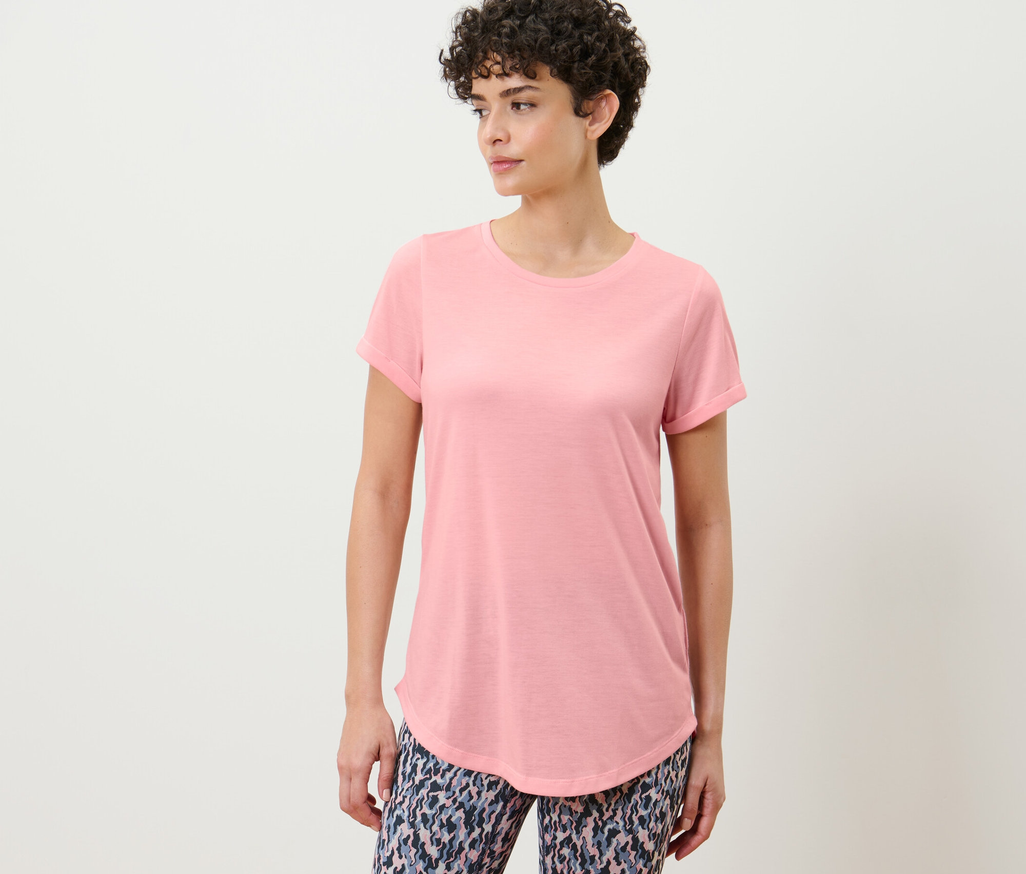 Žena pózuje v dlouhém tričku světle růžové barvy.