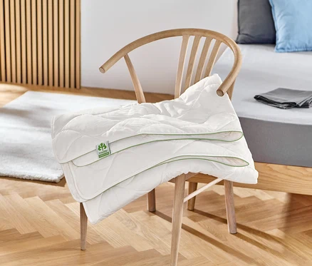 Na dřevěné židli leží složená bílá prošívaná bavlněná přikrývka irisette® greenline.