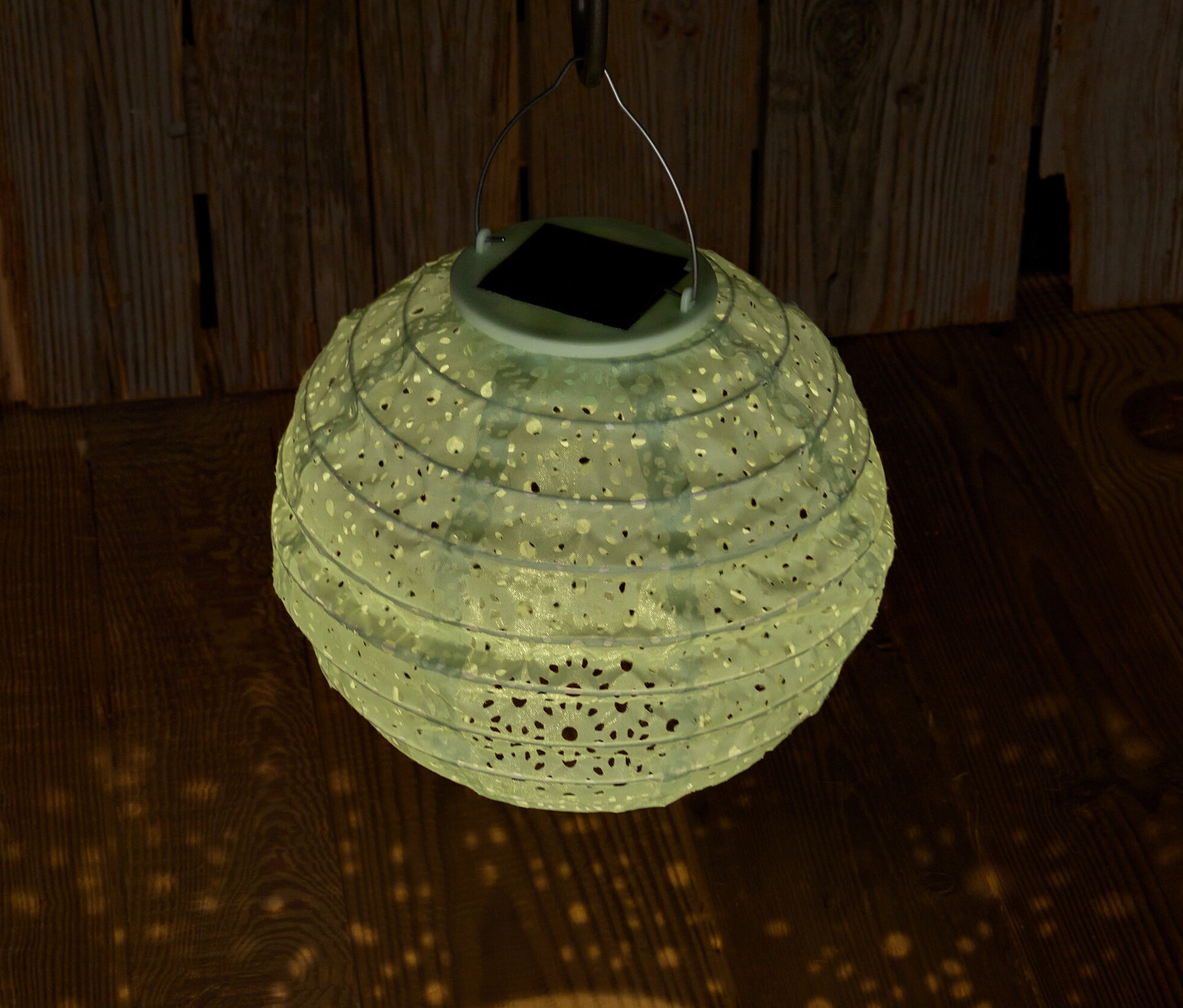Zavěšený solární lampión s ozdobnými otvory promítá světlo na dřevěnou podlahu.