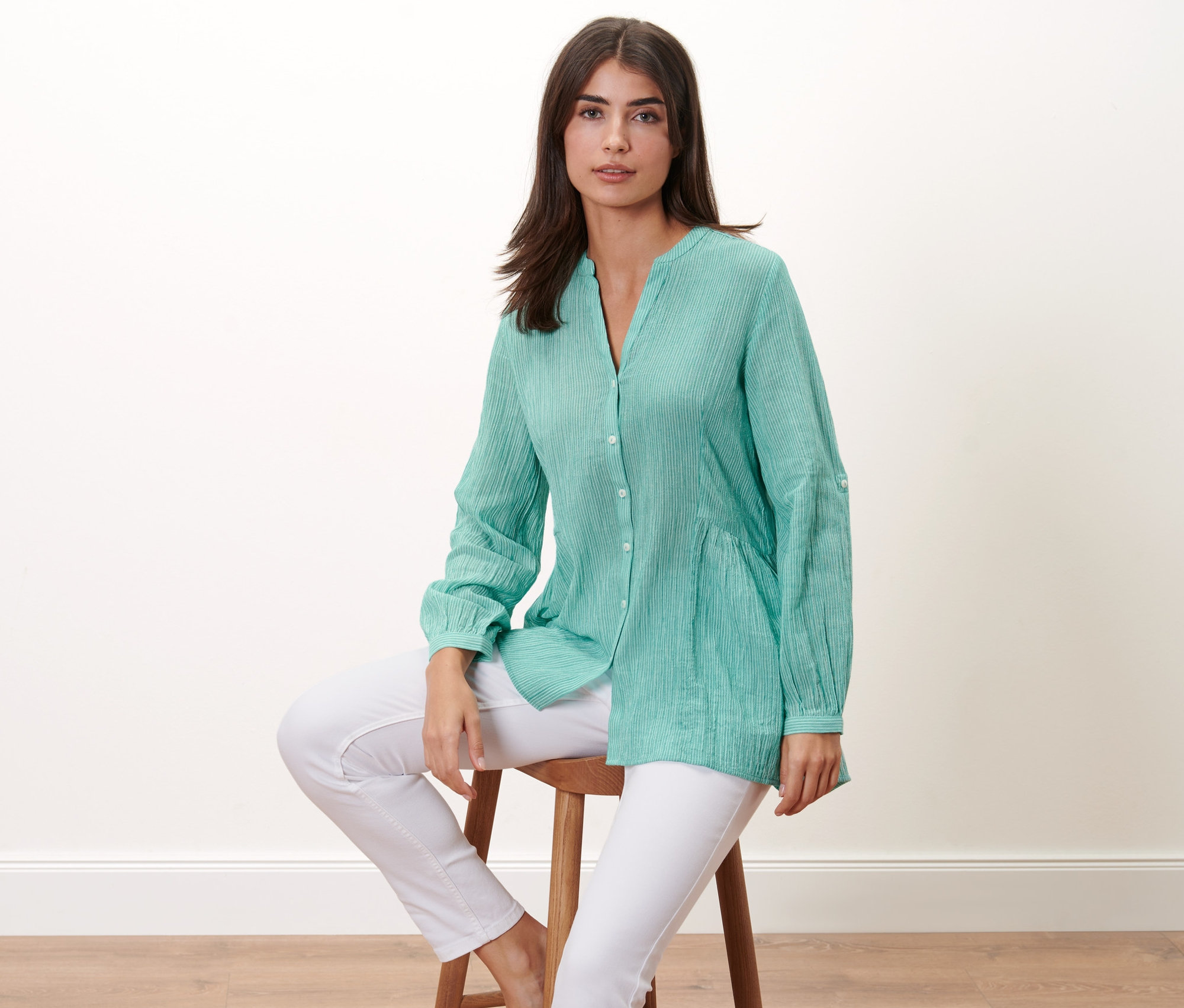 Žena sedí na stoličce a má na sobě proužkovanou dlouhou halenku a bílé kalhoty.