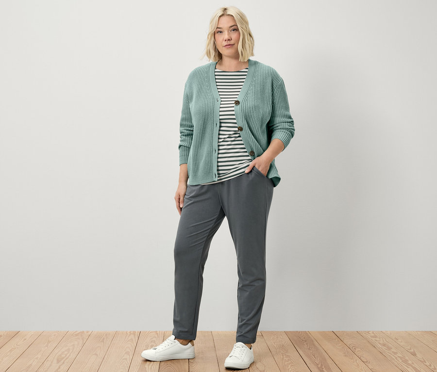 Žena stojí a má na sobě joggpant, kardigan z hrubého úpletu v šalvějové barvě a triko s polodlouhým rukávem se zelenými proužky.