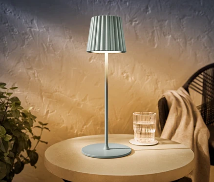 Šalvějově zbarvená LED dobíjecí venkovní stolní lampa stojí na kulatém stolku vedle sklenice vody.