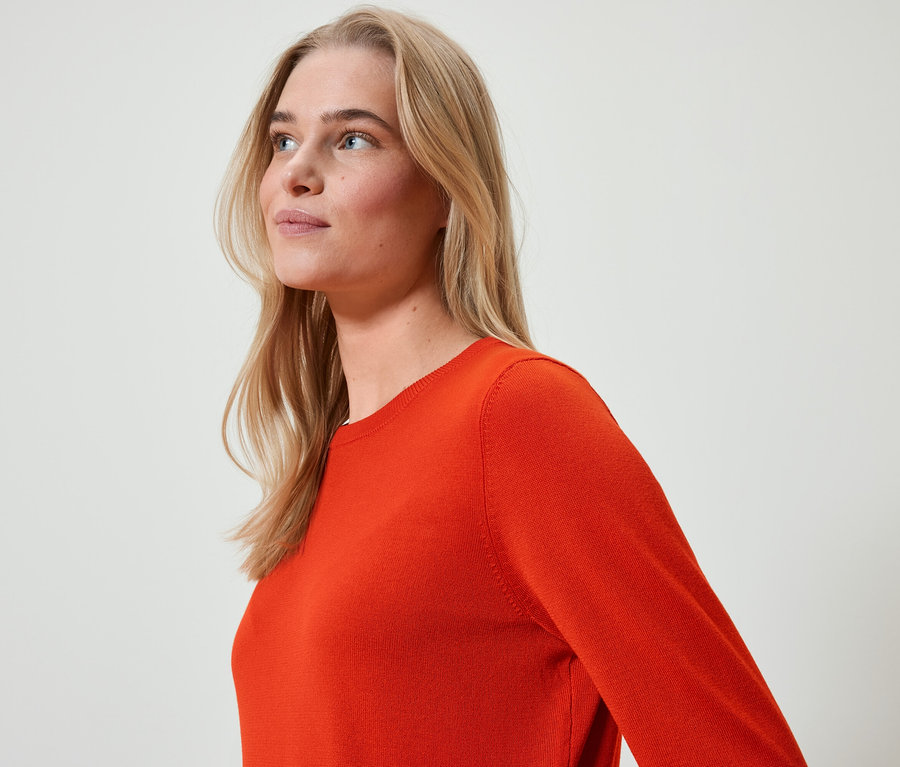 Portrét ženy s blond vlasy v oranžovém svetru, dívající se doleva.