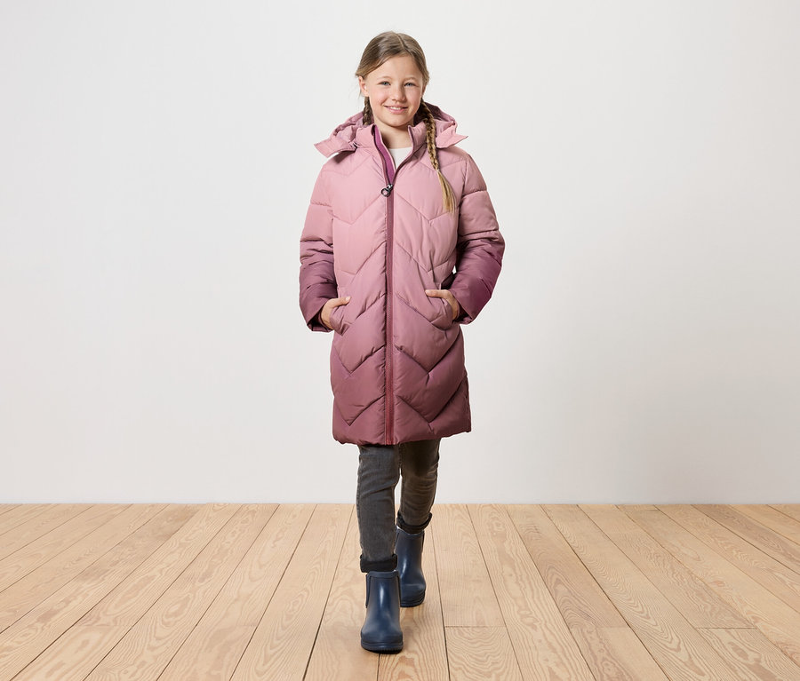 Dívka s blonďatými copy kráčí po dřevěné podlaze a má na sobě dlouhý prošívaný kabát s kapucí, který přechází z růžové do fialové.