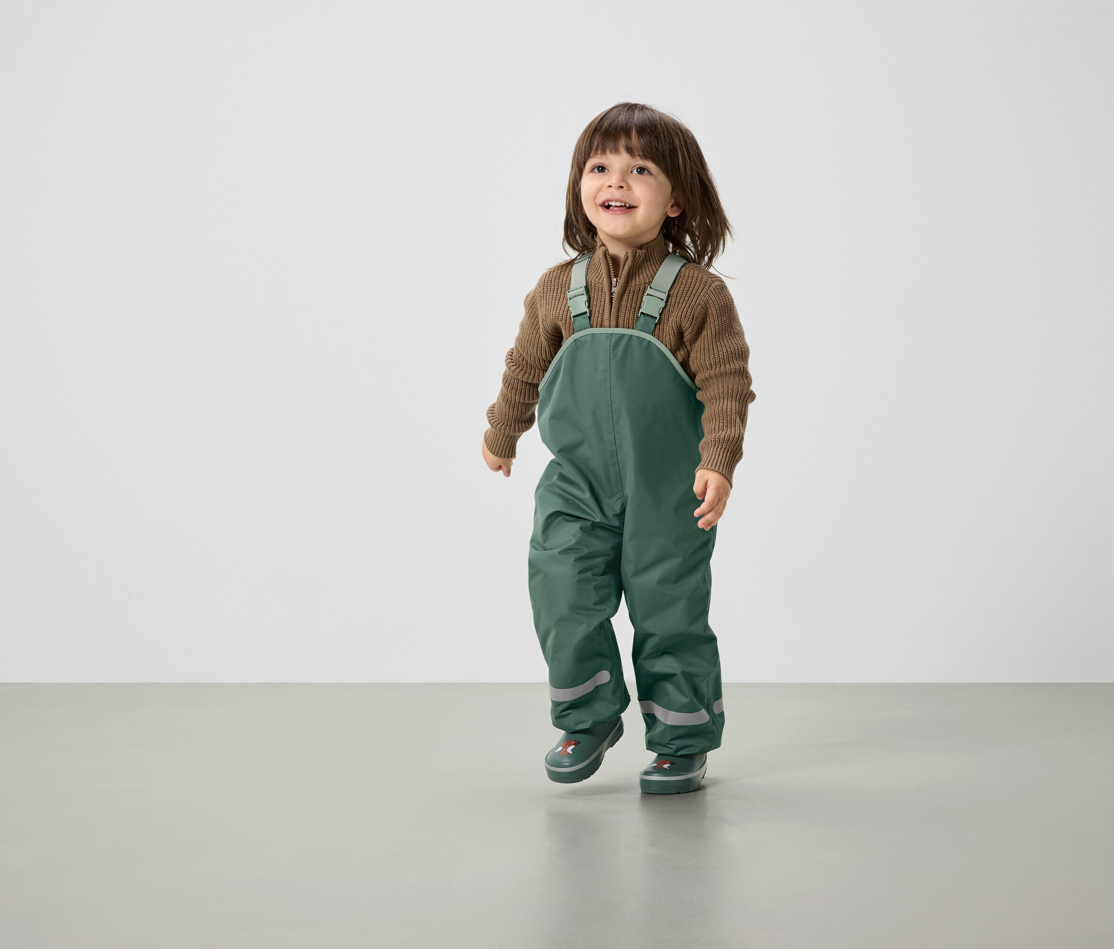 Usmívající se dítě s hnědými vlasy kráčí v zelených kalhotách s laclem a hnědém svetru.
