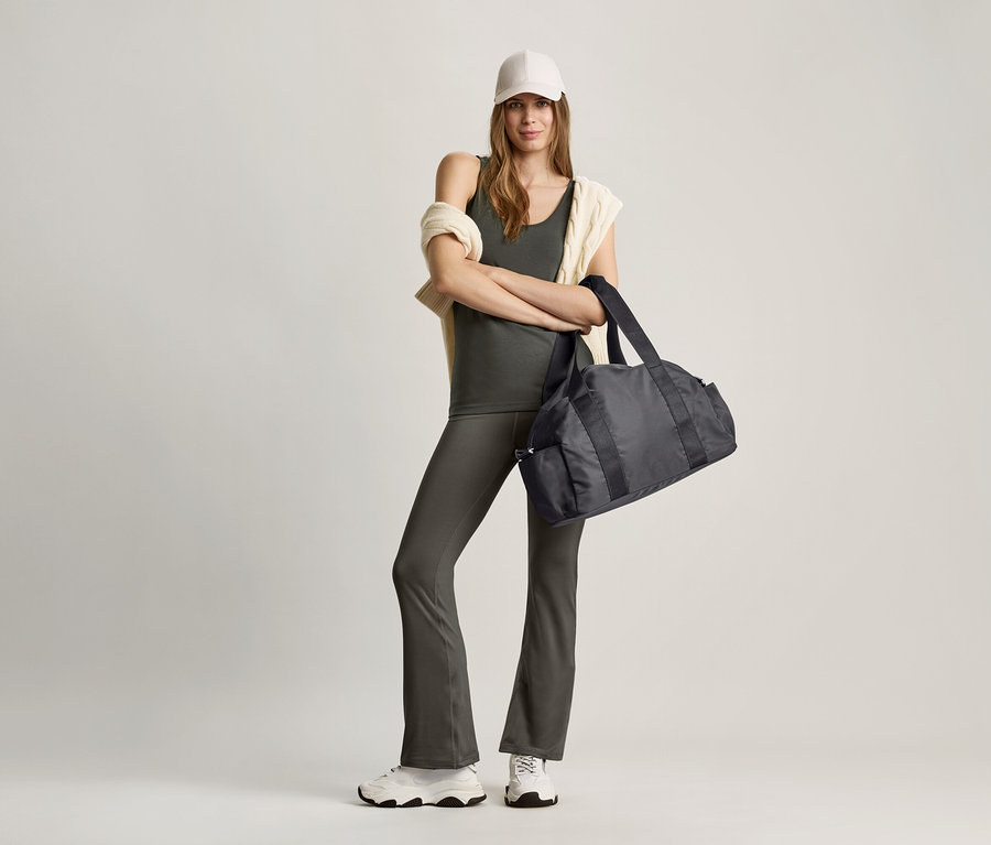 Žena stojí se zkříženýma rukama a drží černou sportovní tašku. Má na sobě olivový top a šedé kalhoty.