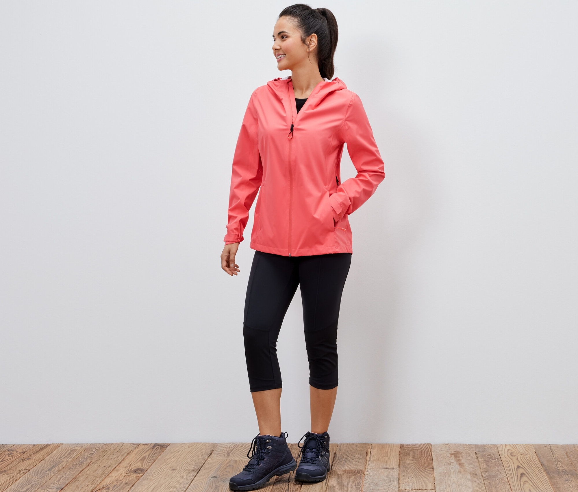 Usmívající se žena pózuje v korálové 2,5vrstvé outdoorové bundě, černých tříčtvrtečních legínách a trekových botách.