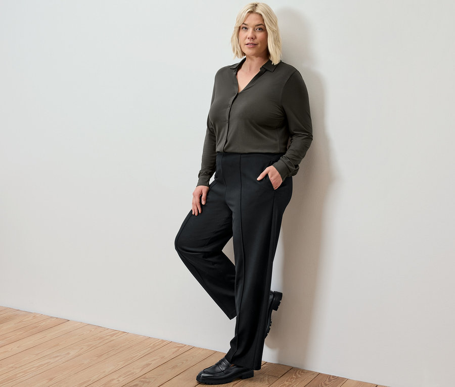 Blonďatá žena opírající se o zeď má na sobě černé žerzejové kalhoty s prošitím a halenkové triko s knoflíkovou légou.