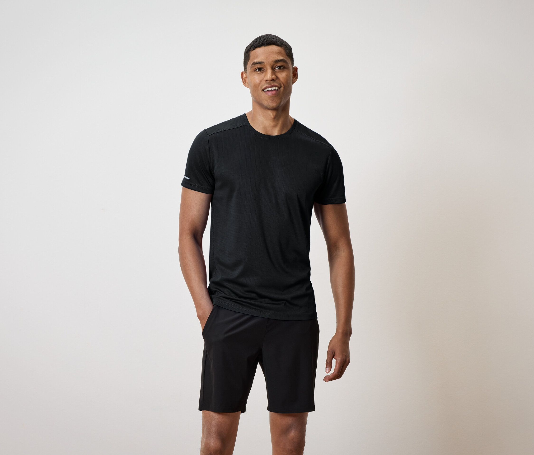 Usmívající se muž stojí a má ruce v kapsách černých šortek a má na sobě černé tričko.