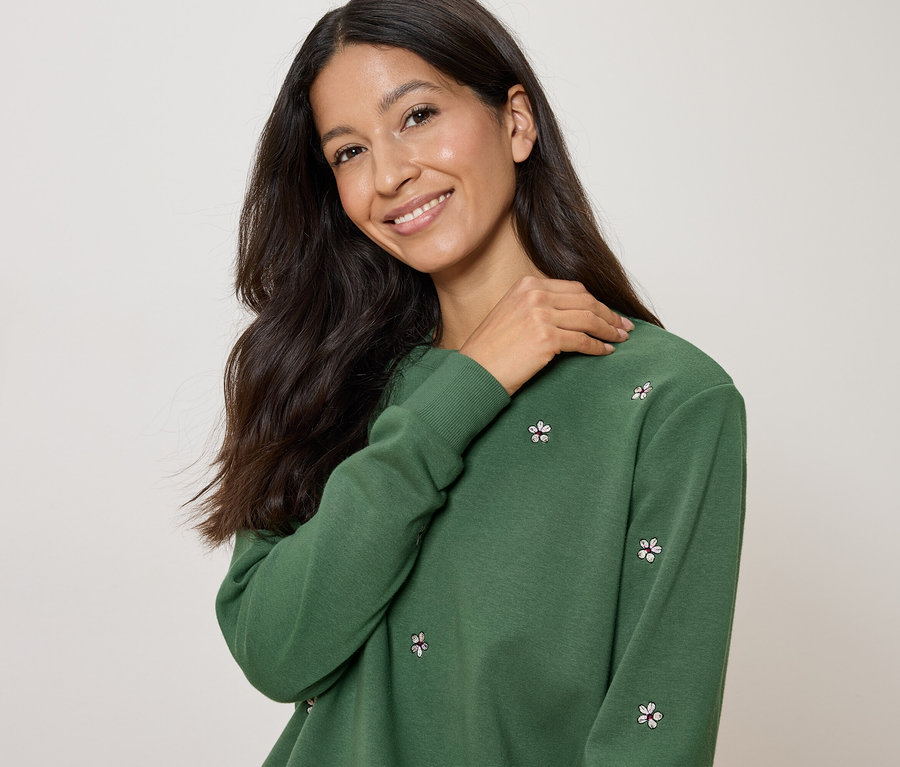 Žena s tmavými, dlouhými vlasy má na sobě zelenou mikinu s květinovým vzorem a klade si ruku na rameno.