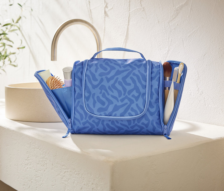 Otevřená modrá kosmetická taška s abstraktním vzorem stojí na bílé desce v koupelně.